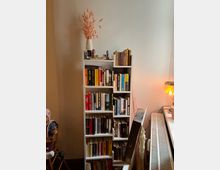 Ein Bücherregal mit mehreren Regalböden voller Bücher verschiedener Größen und Farben steht an einer weißen Wand. Oben auf dem Regal steht eine Dekoration mit getrockneten Blumen in einer weißen Vase, daneben kleinere Dekorartikel; rechts ist ein Heizkörper und eine Tischlampe zu sehen.
