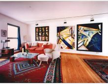 Ein helles Wohnzimmer mit Holzboden, einer roten Couch und zwei weißen Sesseln. An der weißen Wand hängen große, abstrakte Gemälde, und der Raum ist mit orientalischen Teppichen dekoriert.