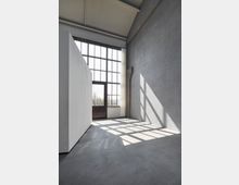 Industrieähnlicher Innenraum mit hohen Decken, großen Fenstern im Gittermuster und glatten Betonwänden. Sonnenlicht fällt durch die Fenster, wirft quadratische Schatten auf den polierten Betonboden.