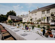 Lange, festlich gedeckte Tafel im Freien mit weißer Tischdecke, Geschirr, Gläsern und Blumenvasen auf einer Rasenfläche. Im Hintergrund steht ein helles Landhaus mit Schieferdach, vielen Fenstern und einer kleinen Turmspitze neben weiteren Nebengebäuden und Bäumen.