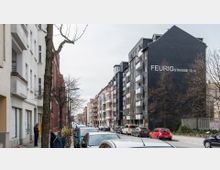 Straßenansicht mit mehrstöckigen Wohngebäuden, darunter ein Gebäude mit schwarzer Fassade und der Beschriftung „Feurigstraße 13, 14“. Parkende Autos säumen die Straße, während kahle Bäume den Gehweg begrenzen.