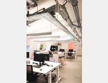 Modernes Büro mit offener Gestaltung, weißen Schreibtischen und mehreren Computerarbeitsplätzen. Sichtbare Lüftungsrohre und Kabeltrassen an der Decke unterstreichen den industriellen Stil, während vereinzelte Dekorationselemente wie Pflanzen und Poster den Raum beleben.