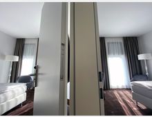 Ein schmaler Durchgang trennt zwei nahezu identische Hotelzimmer mit weißen Wänden, großen Fenstern mit halbtransparenten Vorhängen und dunklen Verdunkelungsgardinen. Die Zimmer enthalten jeweils ein Bett, einen Sessel und eine Stehlampe, mit Teppichboden in dunklen Farbtönen.