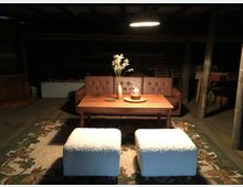 Gemütlicher Wohnbereich in einem rustikalen Dachboden mit Holzboden und sichtbaren Holzbalken. Das Setting umfasst ein braunes Ledersofa, einen Holztisch mit einer Vase und Kerze, zwei flauschige Hocker und einen floral gemusterten Teppich, beleuchtet durch eine Hängelampe.