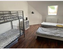 Schlichtes Schlafzimmer mit zwei Einzelbetten und einem Etagenbett, alle mit weiß-blau gemusterten Bettdecken. Der Raum hat einen Holzboden, ein Dachflächenfenster, ein Heizkörper unter dem Fenster, einen offenen Kleiderständer und ein kleines Regal.