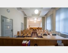 Ein kleiner Gerichtssaal mit hellen Holzmöbeln, einer Reihe hölzerner Besucherstühle, einem erhöhten Richterpult im Hintergrund und weißen Wänden. Große Fenster mit vertikalen Jalousien sowie eine Uhr und Türen an den Seitenwänden prägen den Raum.