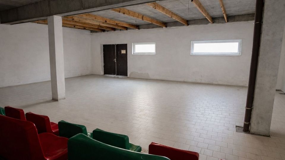 Großer, leerer Innenraum mit gefliestem Boden, weiß verputzten Wänden und Beton-Decke mit sichtbaren Holzbalken. An der Rückwand befinden sich eine schwarze Doppeltür und zwei schmale, horizontale Fenster; im Vordergrund sind Reihen von roten und grünen Kunststoffsitzen zu sehen.