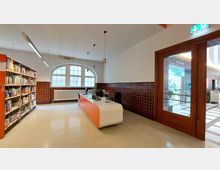 Innenraum einer Bibliothek mit einem Empfangstresen im Vordergrund, einem Bücherregal auf der linken Seite und einem großen, halbrunden Fenster im Hintergrund. Die Wände sind unten mit dunklen Fliesen verkleidet und oben weiß gestrichen; rechts führt eine Holztür mit Glaseinsatz zu einem weiteren Raum.