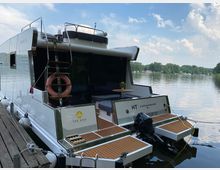 Hausboot mit moderner weißer Konstruktion, vertäut an einem Steg vor einem ruhigen See. Das Boot verfügt über eine Außenterrasse mit Holzboden, Sitzbereich und einem kleinen Tisch, sowie eine kleine Leiter und einen Außenbordmotor. Im Hintergrund sind Bäume und vereinzelte Gebäude am gegenüberliegenden Ufer zu sehen.