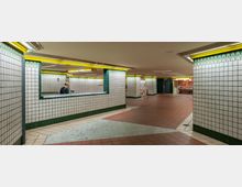 Unterirdischer Durchgang mit weißen Fliesenwänden, grünem Fliesenrand und gelben Beleuchtungselementen. Im Hintergrund befinden sich Shops und ein Informationsschalter, während der Boden mit roten und beigen Fliesen gestaltet ist.