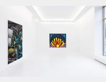 Ein heller, minimalistischer Galerieraum mit weißen Wänden und Böden. Zwei abstrakte Gemälde sind ausgestellt, eines links mit organischen Formen und gedeckten Farben und eines frontal mit leuchtenden Gelb- und Rottönen. Ein großes Fenster rechts lässt Tageslicht herein.