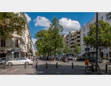 Straßenecke in einer innerstädtischen Umgebung mit mehrstöckigen Wohn- und Geschäftsgebäuden im Altbau- und moderneren Stil. Im Vordergrund stehen Bäume, geparkte Autos, ein Mülleimer und ein Fahrrad; Straßenschilder zeigen "Mommsenstraße" und "Knesebeckstraße".