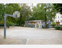 Ein kleiner Basketballplatz mit einem Metallkorb und graffitibeschmierter Säule, umgeben von Bäumen. Im Hintergrund steht ein weißes Gebäude mit bunten Graffiti auf der Fassade, eingebettet in eine parkähnliche Umgebung.