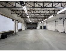 Weitläufiger, industrieller Raum mit sichtbaren Betonwänden und freiliegender Stahlkonstruktion an der Decke. Der Raum ist mit Scheinwerfern auf Stativen, weißen Projektionsflächen an den Wänden und geschlossenen Türen ausgestattet.