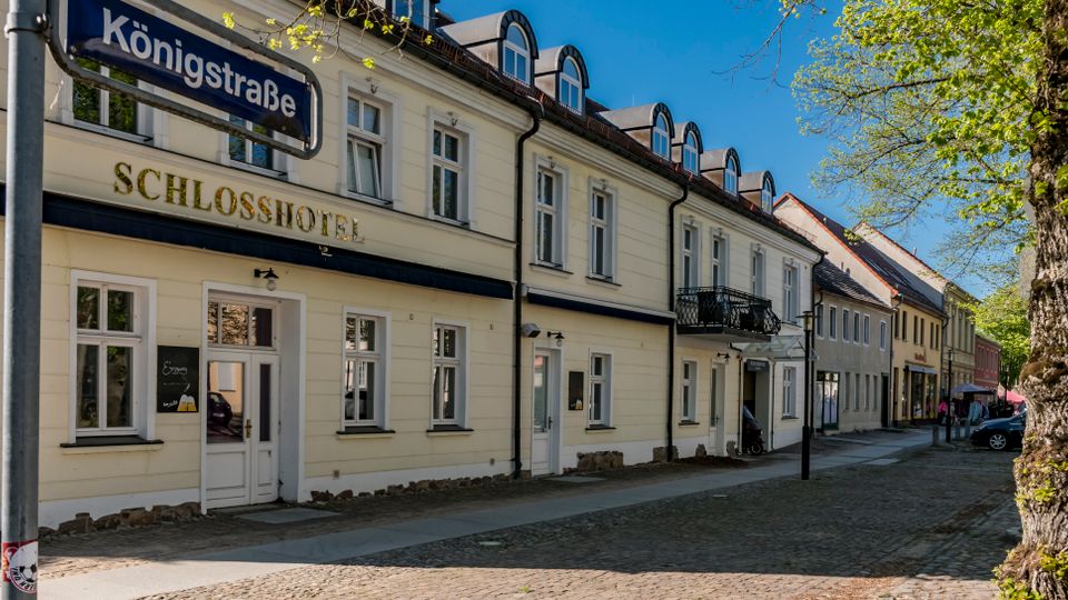 Street view of a cream-colored historic hotel building labeled “Schlosshotel” along Königstraße, with tall windows, dormer windows on the roof, and a small balcony. A cobblestone sidewalk runs beside a tree-lined curb with parked cars under a clear blue sky.
