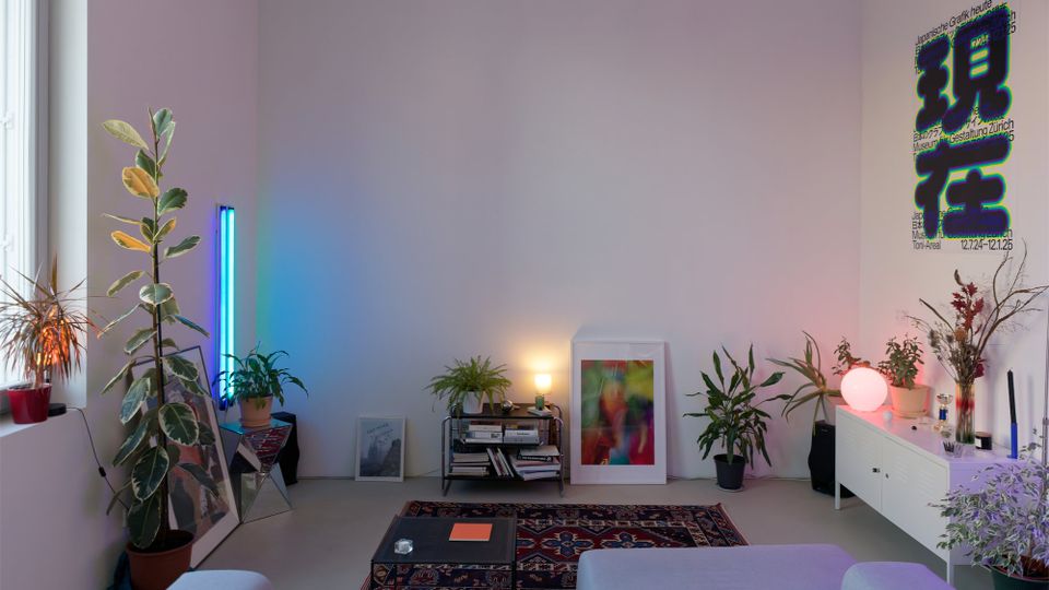 Modernes Wohnzimmer mit großer weißer Wand, vielen Zimmerpflanzen und indirekter Beleuchtung durch eine blaue LED-Röhre und eine runde Tischlampe. Vor der Wand stehen ein niedriges Regal mit Büchern, gerahmte Bilder am Boden, ein gemusterter Teppich mit Couchtisch sowie ein Sideboard mit Deko und Poster.
