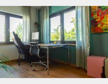 Modernes, helles Homeoffice mit einem Schreibtisch aus Metall und Holz, einem schwarzen Bürostuhl und einem Computerbildschirm. Große Fenster mit weißen Vorhängen bieten Blick ins Grüne, während ein buntes abstraktes Kunstwerk an der türkisfarbenen Wand und ein rosa Beistelltisch den Raum ergänzen.