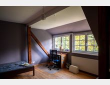 Dachgeschosszimmer mit schräger Decke, Holzboden und sichtbarem Holzbalken, eingerichtet mit Bett und kleinem Schreibtisch. Drei große Sprossenfenster lassen Tageslicht herein und zeigen Bäume draußen.