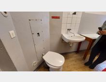 Kleines Badezimmer mit einfacher Ausstattung: ein weißes Waschbecken, eine Toilette, helle Wandfliesen im Bereich des Waschbeckens und graue Wände. Der Boden besteht aus hellem Laminat, und in der Ecke steht ein Holztisch.