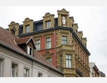 Mehrstöckiges Gebäude in historistischem Stil mit gelb-ockerfarbenen und ziegelroten Fassadenelementen sowie grünen Fensterrahmen. Das Dach ist mit Gauben und einer Statue dekoriert, im Vordergrund sind Teile benachbarter Häuser mit Ziegeldächern sichtbar.