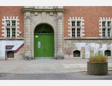 Backsteinfassade eines Gebäudes mit einem großen grünen Eingangstor, flankiert von steinernen Pilastern und Fenstern mit weißen Rahmungen. Der untere Bereich der Fassade ist teils mit Graffiti bedeckt, und im Vordergrund steht ein runder Betonpflanzkübel mit Grasbewuchs.