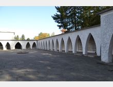 Ein rechteckiger Innenhof mit einem gepflasterten Boden wird von einem Arkadengang aus weiß verputzten Bögen umgeben. Im Hintergrund sind hohe Bäume und ein blauer Himmel zu sehen.