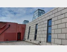 Flachdachbereich mit einer roten Wand und einer beigen Fassade, in die mehrere vertikale Fenster eingebaut sind. Im Hintergrund sind gläserne pyramidenförmige Lichtkuppeln sichtbar, der Himmel ist leicht bewölkt.