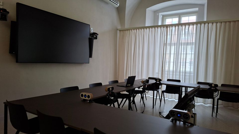 Besprechungsraum mit U-förmig angeordneten dunklen Tischen und Stühlen, ausgestattet mit mehreren Kameras, Mikrofonen und einem Laptop für Videokonferenzen. An der Wand hängt ein großer Bildschirm; gegenüber befindet sich ein großes Fenster mit halbtransparenten Vorhängen.