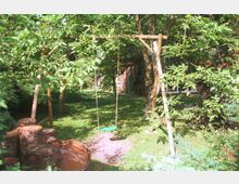 Ein Garten mit dichtem grünen Laub, einer Holzschaukel mit grünem Sitz und einem Stapel aus gesägten Baumstämmen. Im Hintergrund ist ein Haus mit roten Ziegeln und Fensterfronten zu sehen.
