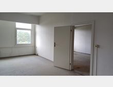 Ein leerer Raum mit weißen Wänden und einem einfachen Fenster, das Blick auf Bäume bietet. Eine offene Tür führt in einen angrenzenden, ebenfalls leeren Raum mit ähnlicher Ausstattung.