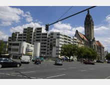 Straßenszene mit Kreuzung, im Hintergrund ein modernes Wohn- und Geschäftshaus mit Balkonen auf der linken Seite und ein historisches Gebäude mit einem hohen Turm und roten Dächern auf der rechten Seite. Im Vordergrund fahren Autos über die Straße, während Bäume den Gehweg säumen.