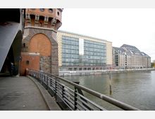 Blick auf einen Abschnitt der Oberbaumbrücke in Berlin mit rotem Backsteinturm im Vordergrund, daneben eine moderne Gebäudefassade aus Glas und hellem Stein am Ufer der Spree. Im Vordergrund verläuft ein Geländer entlang eines Gehwegs.
