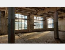 Ehemaliger Fabrikraum mit großen, vergitterten Fenstern, durch die Tageslicht einfällt. Der Raum hat sichtbare Alterungsspuren, mit abgeblätterter Wandfarbe, Holzboden und freiliegenden Rohrleitungen an der Decke.