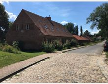 Ein niedriger, länglicher Backsteinbau mit rotem Satteldach und mehreren Dachfenstern, umgeben von Blumenbeeten und einer Grünfläche, steht an einer gepflasterten Dorfstraße. Im Hintergrund sind weitere Backsteinhäuser und Bäume zu sehen, bei sonnigem Wetter.