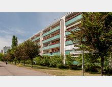 Mehrstöckiges Wohngebäude mit grünen Balkonen und roten Blumenkästen, umgeben von Bäumen und einem gepflegten Gehweg mit Sitzbänken. Ein Straßenschild weist auf den "Mühlenbecker Weg".