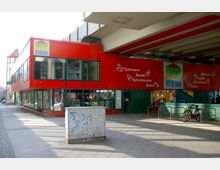 Ein einstöckiges Gebäude mit roter Fassade, großen Glasfenstern und der Aufschrift "Werken, Spielen, Schenken". Darunter befinden sich Geschäfte für Spielwaren, Bastelbedarf, Schreibwaren und ein Bistro; im Vordergrund steht ein Stromkasten und es gibt einen Fahrradständer.