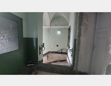 Treppenhaus in einem älteren Gebäude mit abgeblätterter Wandfarbe und einem Marmor-Treppengeländer. Im Hintergrund ein beleuchtetes Notausgangsschild und eine gewölbte Decke.