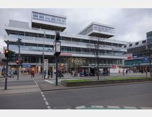 Eine Straßenansicht des Einkaufszentrums "Forum Steglitz" in Berlin. Das moderne Gebäude hat eine gläserne Fassade im Erdgeschoss mit Geschäften wie Rossmann und Apollo sowie darüber liegende Büroetagen mit großen Fensterfronten; davor befindet sich eine belebte Straße mit Ampeln und Fußgängern.