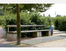 Ein Park mit einer einfachen Holzbank auf Steinblöcken, neben einem Baum und umgeben von dichtem grünen Gebüsch. Im Hintergrund ist der Berliner Fernsehturm sichtbar.