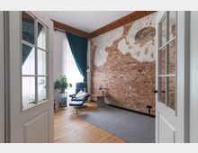 Ein gemütliches Zimmer mit einer freiliegenden Ziegelwand, teilweise verputzt, und zwei hohen Fenstern mit dunkelgrünen Vorhängen. Im Raum befinden sich ein schwarzer Ledersessel mit Fußhocker, ein kleiner Holztisch, eine Zimmerpflanze und ein heller Holzboden mit einem grauen Teppich.