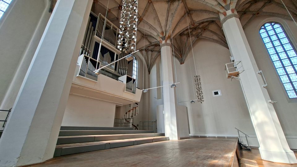 Innenraum einer hohen Kirche mit Rippengewölbe, weißen Pfeilern und großen Spitzbogenfenstern. Links sind eine erhöhte Orgelempore mit sichtbaren Orgelpfeifen und darunter ansteigende Sitzstufen sowie eine freie Bühne im Vordergrund zu sehen.