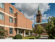 Rotes Backsteingebäude mit großen Fenstern links im Vordergrund und einer Kirche aus Backstein mit grünem Turmdach im Hintergrund. Im Vordergrund befinden sich Büsche, ein Gehweg und ein kleiner Parkplatz, bei bewölktem Himmel.