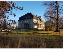 Ein verlassenes Herrenhaus mit verwitterter Fassade und mehreren Etagen steht inmitten einer herbstlichen Parklandschaft mit kahlen Bäumen und verstreutem Laub. Im Vordergrund befinden sich Gras, Büsche und ein Zaun, während der Hintergrund blauen Himmel zeigt.