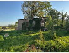 Backsteingebäude mit großen, bogenförmigen Fenstern, dessen Fassade teilweise von Efeu überwachsen ist, inmitten einer ländlichen Wiese. Im Vordergrund liegen hohes Gras und Wildblumen, darüber sind Lichterketten gespannt, umgeben von Bäumen und offenem Himmel.