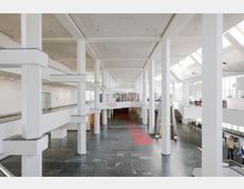 Großer, heller Innenraum mit weiß gestrichenen Säulen und hohen Decken, modernen Treppen, einer Galerie im oberen Bereich und einer Garderobe im Hintergrund. Der Boden besteht aus dunklen Steinfliesen, und durch die Oberlichter fällt viel Tageslicht hinein.