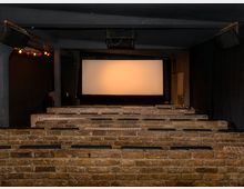 Ein kleiner Kinosaal mit gemauerten Sitzreihen aus Ziegelsteinen und einem beleuchteten weißen Bildschirm im Hintergrund. Dunkle Wände und abgehängte Lautsprecher prägen die minimalistische Einrichtung.