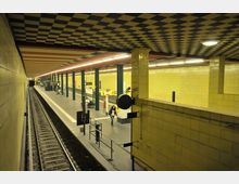 U-Bahn-Station mit gelben gefliesten Wänden, einem schwarz-weiß karierten Deckendesign und zwei gegenüberliegenden Bahnsteigen. Im Vordergrund sind Gleise, Signalanlagen und eine begrenzte Rohrstruktur zu sehen, während vereinzelt Personen auf den Bahnsteigen warten.