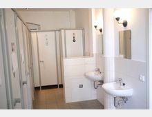 Ein schlicht gestalteter Sanitärraum mit einzelnen Kabinen auf der linken Seite, ausgestattet mit weißen Türen und kleinen Beschilderungen. Rechts befinden sich zwei weiße Waschbecken mit Spiegeln darüber und Wandlampen zur Beleuchtung.