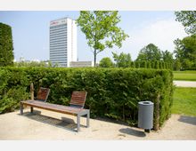 Parklandschaft mit einer Bank aus Holz und Metall vor einer niedrigen Hecke; im Hintergrund ein modernes Hochhaus mit sichtbarem Logo sowie weitere Grünflächen und Bäume.