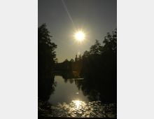 Ein See in einer parkähnlichen Umgebung bei Sonnenuntergang, mit Bäumen am Ufer und der spiegelnden Sonne auf der Wasseroberfläche. Im Hintergrund ist die Silhouette eines Kirchturms zu erkennen.
